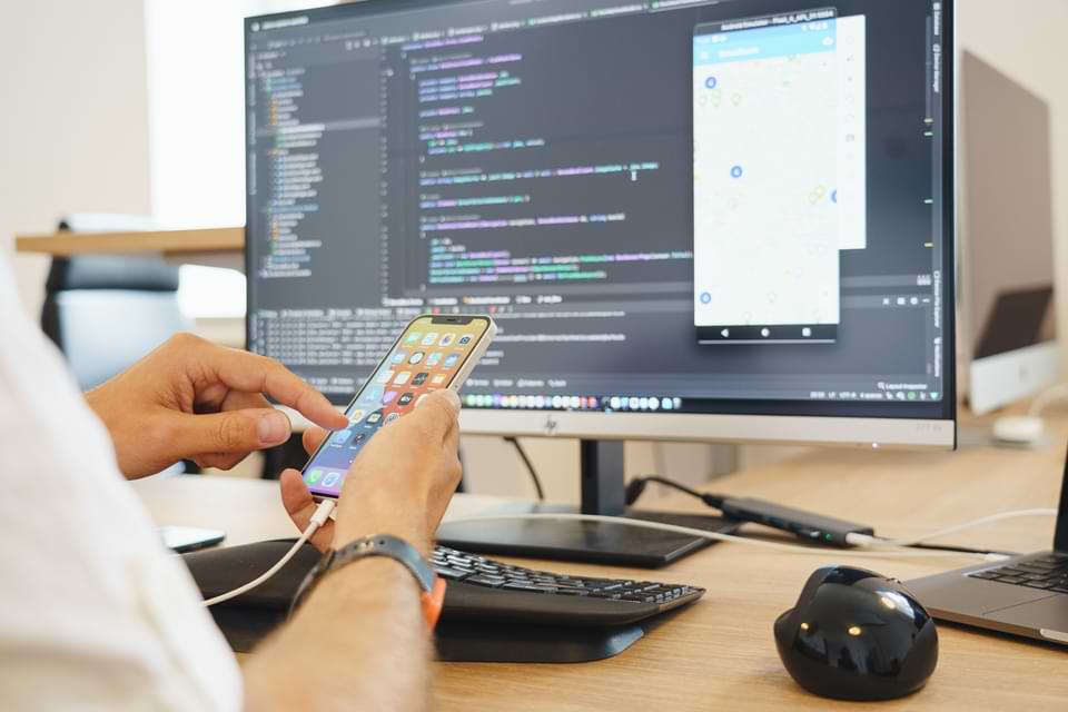 Entwickler testet mobile App auf Smartphone während der App-Programmierung am Desktop – Beispiel für App-Entwicklung und UI/UX-Design bei Cayas.